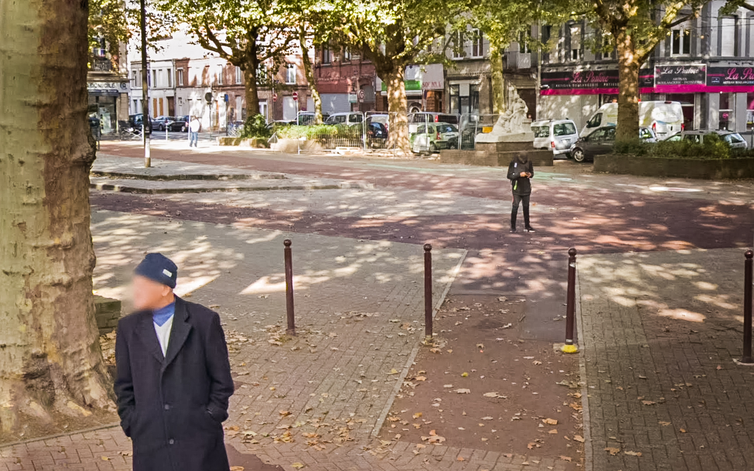 Place Vanhoenacker à Lille, un homme agé au premier plan attend pour traverser. Un jeune homme au second plan regarde son téléphone